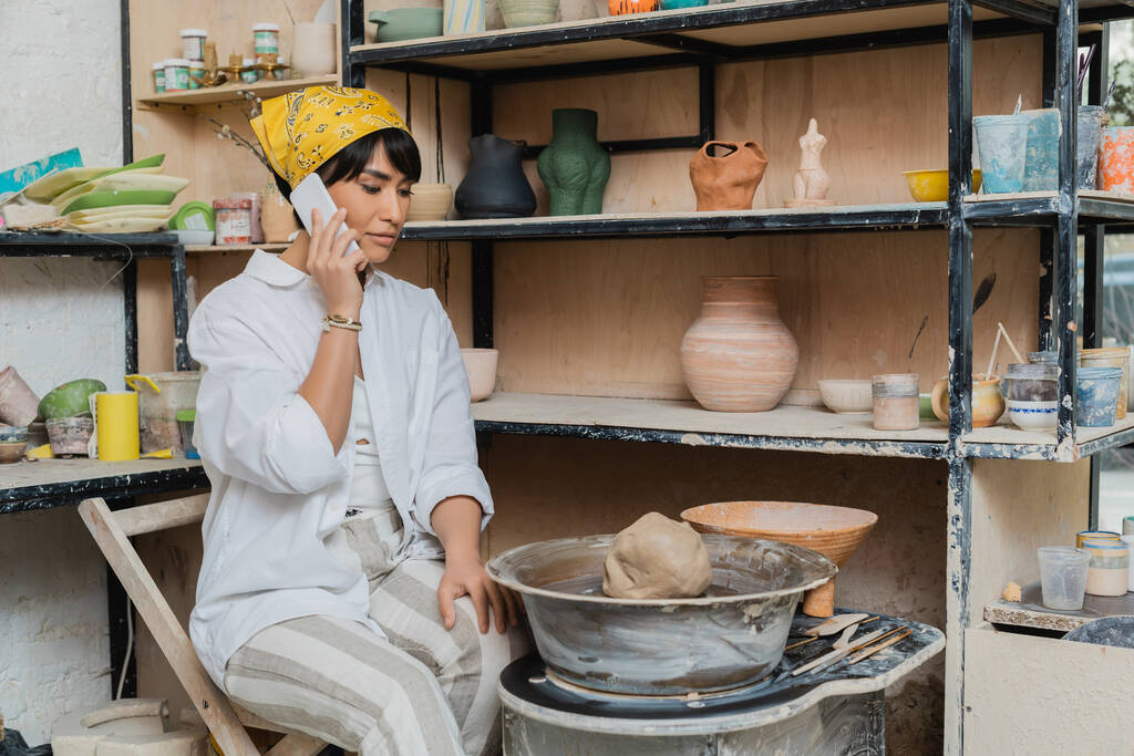Young asian female potter in workwear and headscarf talking on smartphone while sitting near clay on pottery wheel and tools and working in pottery studio, artisan crafting ceramics in studio - Photo, Image