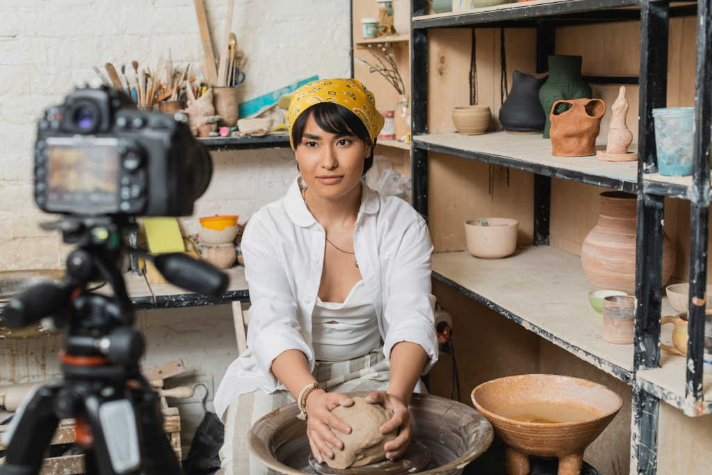 Fiatal barna ázsiai kézműves nő fejkendővel és munkaruha penész agyag kerámia kerék közelében elmosódott digitális fényképezőgép állvány és állvány szobrok stúdióban, fazekas művész bemutató kézműves - Fotó, kép