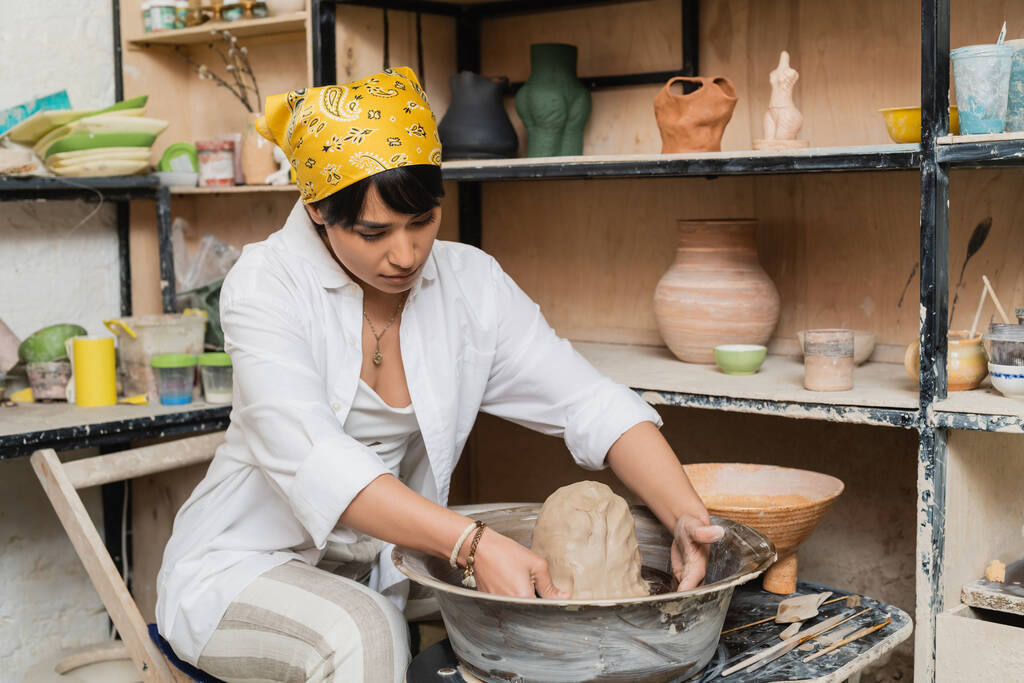 Giovane asiatica donna artigiana in abbigliamento da lavoro e velo stampaggio argilla sulla ruota ceramica vicino a sculture su rack e strumenti in laboratorio di ceramica, ceramica artista in mostra mestiere - Foto, immagini