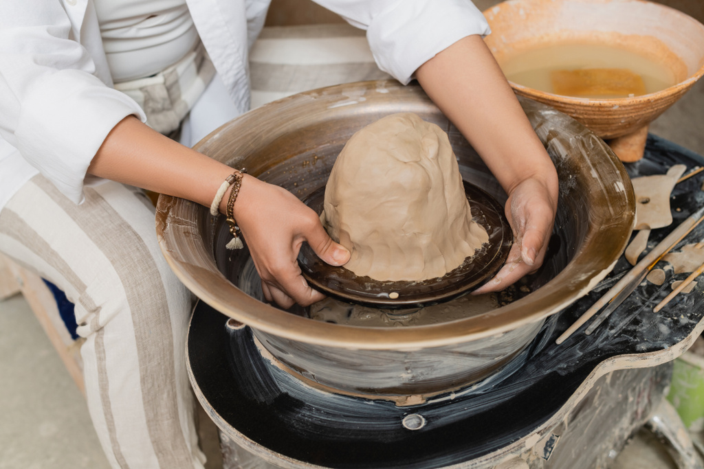 Cropped view of young female artisan in workwear working with clay on pottery wheel, tools and bowl with water at background in ceramic workshop, pottery artist showcasing craft - Photo, Image