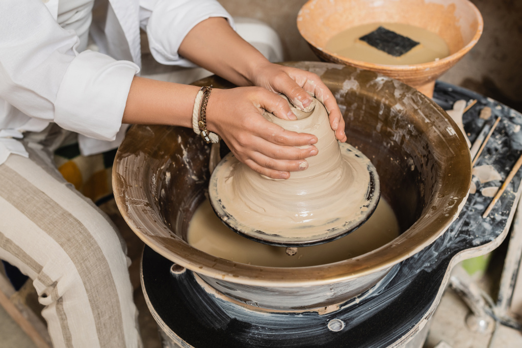 Widok młody rzemieślnik w odzieży roboczej kształtowania mokrej gliny podczas pracy na kole ceramiki w pobliżu zamazanej miski z wodą i gąbką na tle, ceramiki studio przestrzeń robocza i rzemiosła koncepcja - Zdjęcie, obraz