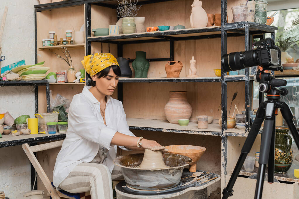 Jeune artisan asiatique en foulard moulant de l'argile humide sur une roue de poterie près d'un appareil photo numérique sur trépied dans un atelier de céramique, un atelier de poterie et un concept artisanal - Photo, image