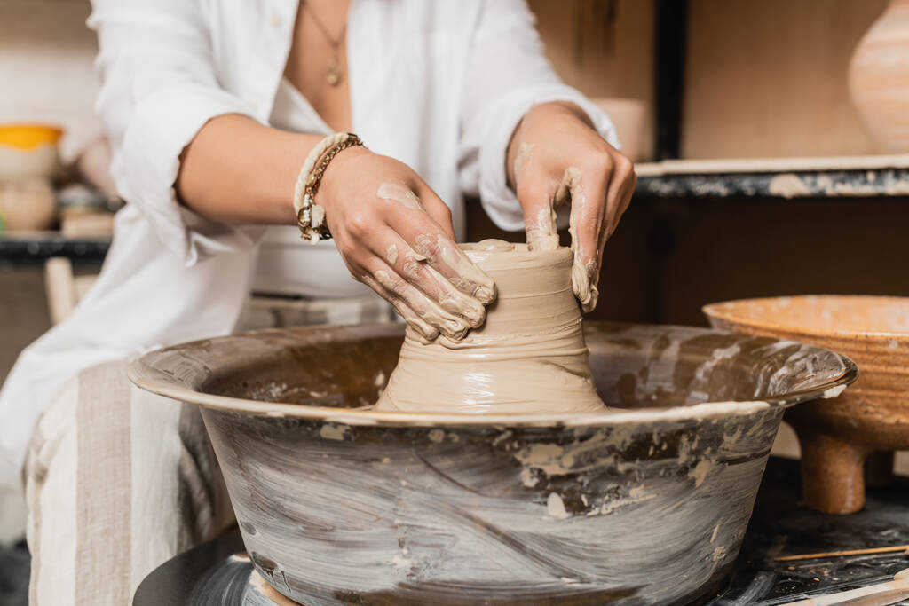 Widok młodych kobiet rzemieślnik w odzieży roboczej kształtowania mokrej gliny podczas pracy na koło ceramiki w pobliżu miski na tle, ceramiki studio obszaru roboczego i koncepcji rzemiosła - Zdjęcie, obraz