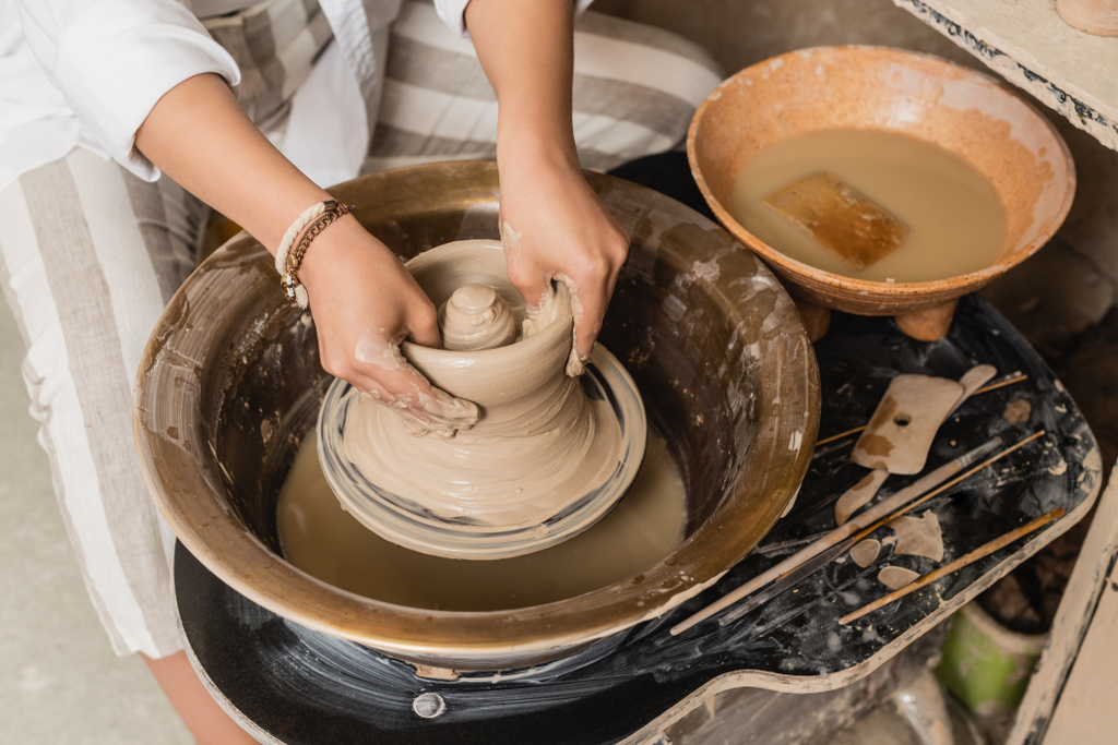 Вид сверху молодой художницы, лепящей мокрые когти на гончарном круге рядом с инструментами и чашей с водой в керамической мастерской, мастерской гончарного дела и концепцией ремесла - Фото, изображение