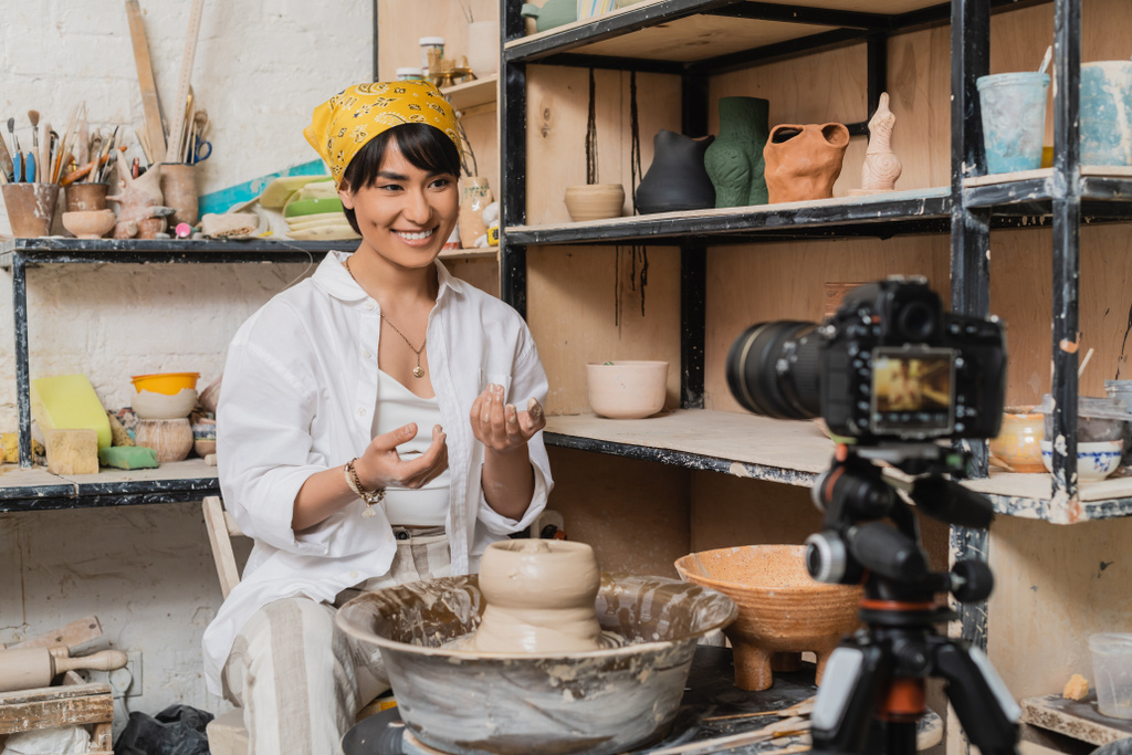 Positif jeune artisan asiatique en foulard et vêtements de travail en regardant l'appareil photo numérique floue sur trépied près de l'argile sur roue de poterie et rack avec des sculptures en atelier, concept de processus de sculpture d'argile - Photo, image
