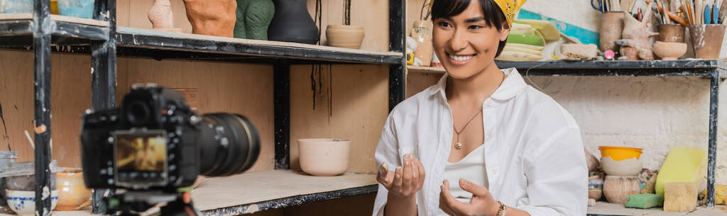 Jovem alegre asiático oleiro feminino falando com câmera digital borrada enquanto sentado perto de esculturas de argila no rack em oficina de cerâmica, conceito de processo de escultura de argila, banner - Foto, Imagem