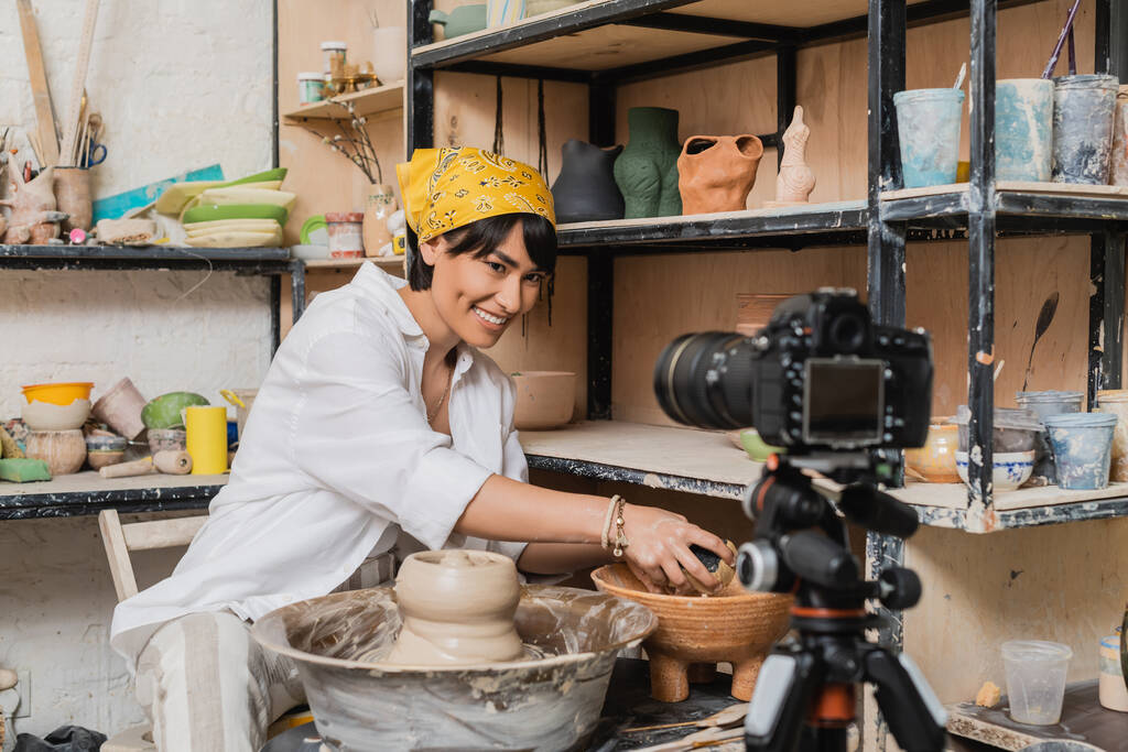 Улыбающаяся азиатская ремесленница в платке, держащая губку возле чаши с водой, глиной и гончарным колесом и глядя на цифровую камеру в художественной мастерской, концепция процесса глиняной скульптуры - Фото, изображение