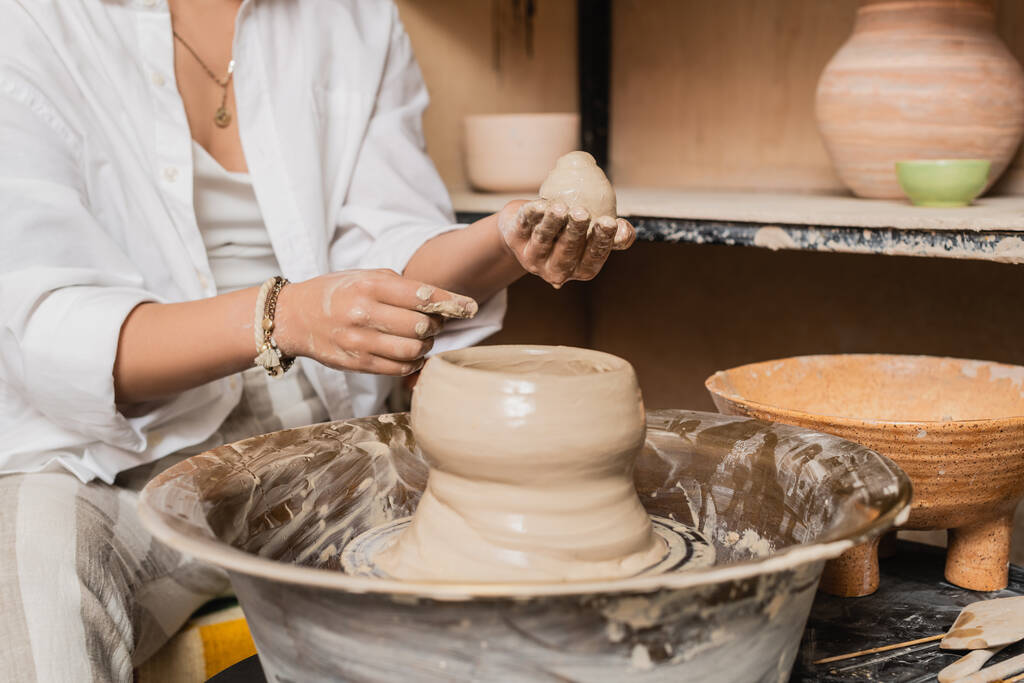 Vista ritagliata di giovane artigiana femminile in azienda abbigliamento da lavoro e stampaggio argilla sulla ruota ceramica e lavorare vicino ciotola in laboratorio di ceramica, concetto di processo di scultura argilla - Foto, immagini