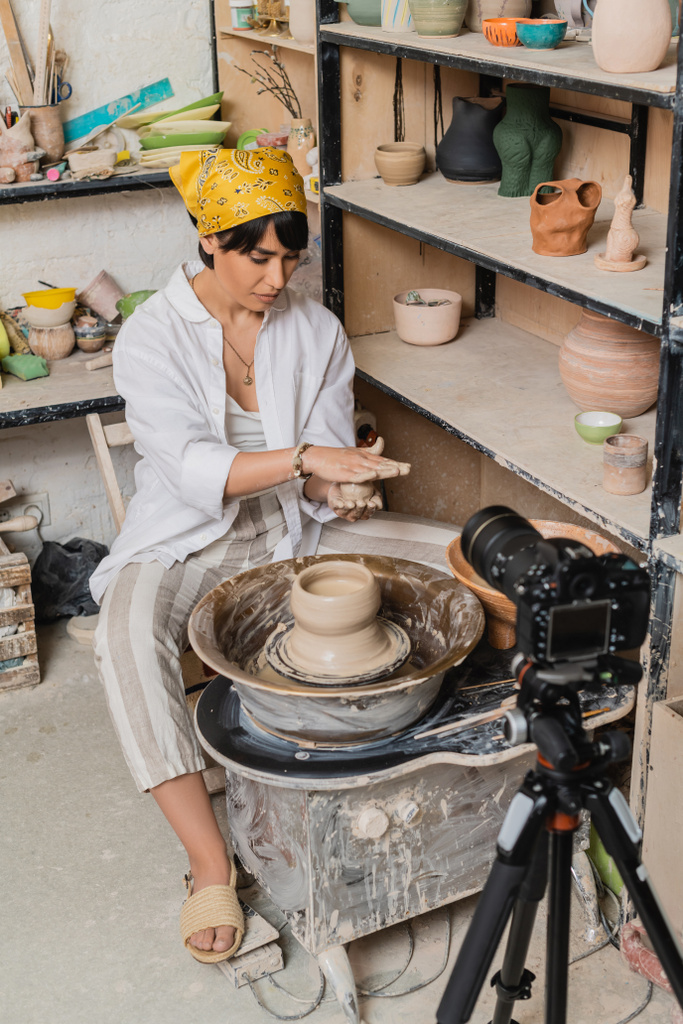 Junge asiatische Kunsthandwerkerin in Kopftuch und Arbeitskleidung hält feuchten Ton in der Hand und arbeitet in der Nähe von Töpferscheibe und Digitalkamera auf Stativ im Keramik-Atelier, Konzept zur Tonbildhauerei - Foto, Bild