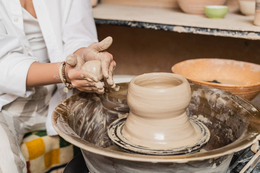Vista ritagliata di giovane ceramista femminile in abbigliamento da lavoro che tiene argilla umida vicino alla ruota della ceramica e ciotola sfocata in studio di ceramica sfocata, concetto di processo di scultura in argilla - Foto, immagini