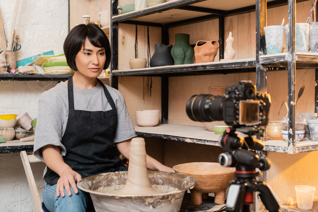 Young asian brunette craftswoman in apron looking at blurred digital camera on tripod near wet clay on pottery wheel and rack in ceramic workshop, pottery tools and equipment - Photo, Image