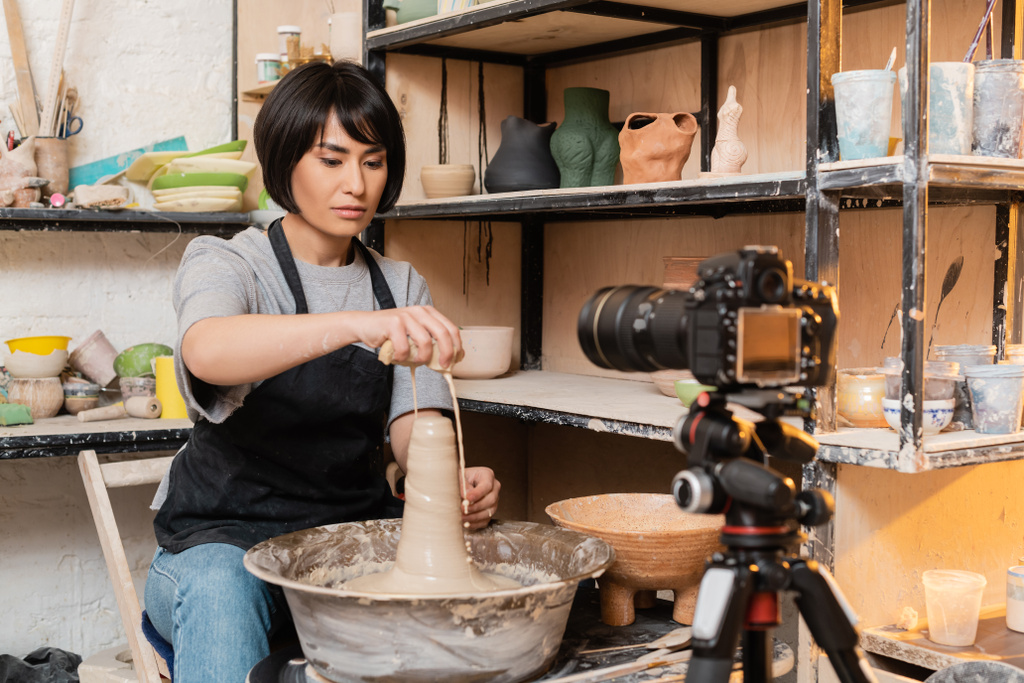 Jovem morena asiática feminina artesã em avental derramando água de esponja em barro molhado e roda de cerâmica perto de câmera digital em tripé em estúdio no fundo, ferramentas de cerâmica e equipamentos - Foto, Imagem
