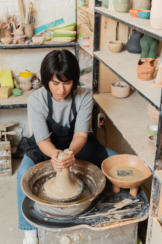 Joven mujer asiática artesana en delantal moldeado arcilla mojada en la rueda de cerámica cerca de cuenco con agua y herramientas mientras trabaja en taller de cerámica, herramientas de cerámica y equipo - Foto, Imagen