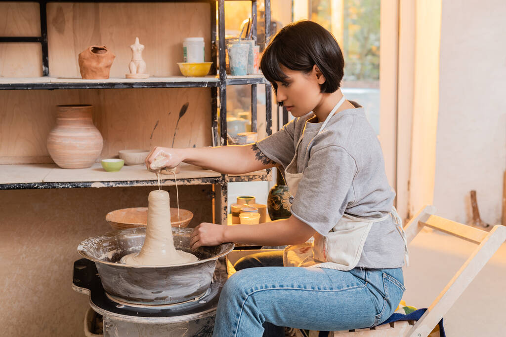陶器の車輪の上の湿った粘土にスポンジから水を注ぐエプロンで若いアジアの女性職人の側面図や日没時のセラミックワークショップで働く、陶器のツールや機器 - 写真・画像