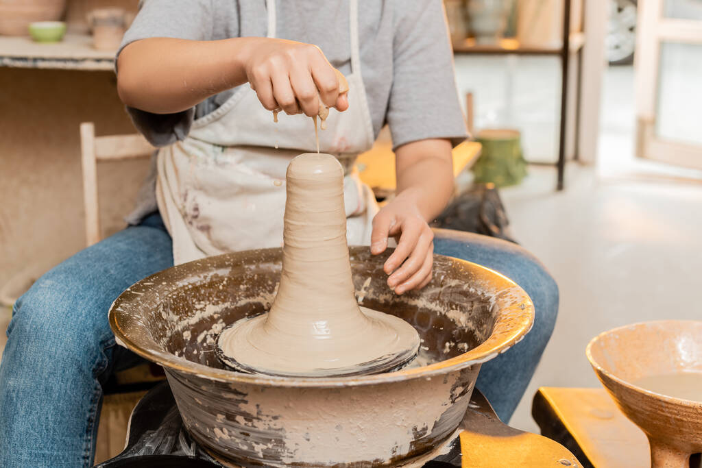 Ausgeschnittene Ansicht einer Kunsthandwerkerin in Schürze, die Wasser auf Ton gießt, während sie an der Töpferscheibe in einer verschwommenen Keramikwerkstatt im Hintergrund arbeitet, Kunsthandwerkerin, die einzigartige Keramikstücke schafft - Foto, Bild