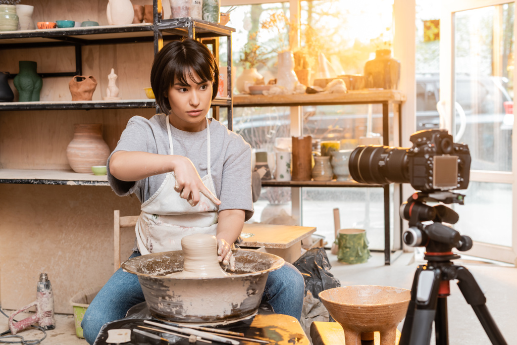 Young asian female potter in apron pointing with finger while working with clay on pottery wheel and looking at digital camera in workshop at sunset, artisan creating unique pottery pieces - Photo, Image