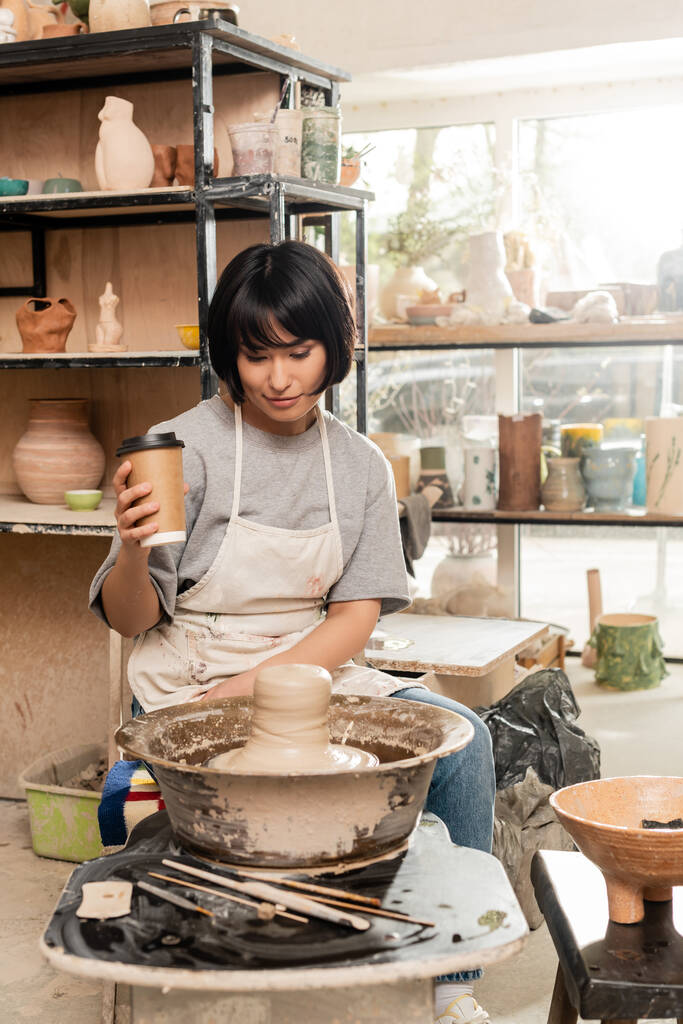 Önlüklü genç Asyalı kadın çömlekçi, çanak çömlek çarkının yakınındaki aletlere bakmak için kahve tutuyor. Bulanık çömlek atölyesinde çanak çömlek yapıyor. - Fotoğraf, Görsel