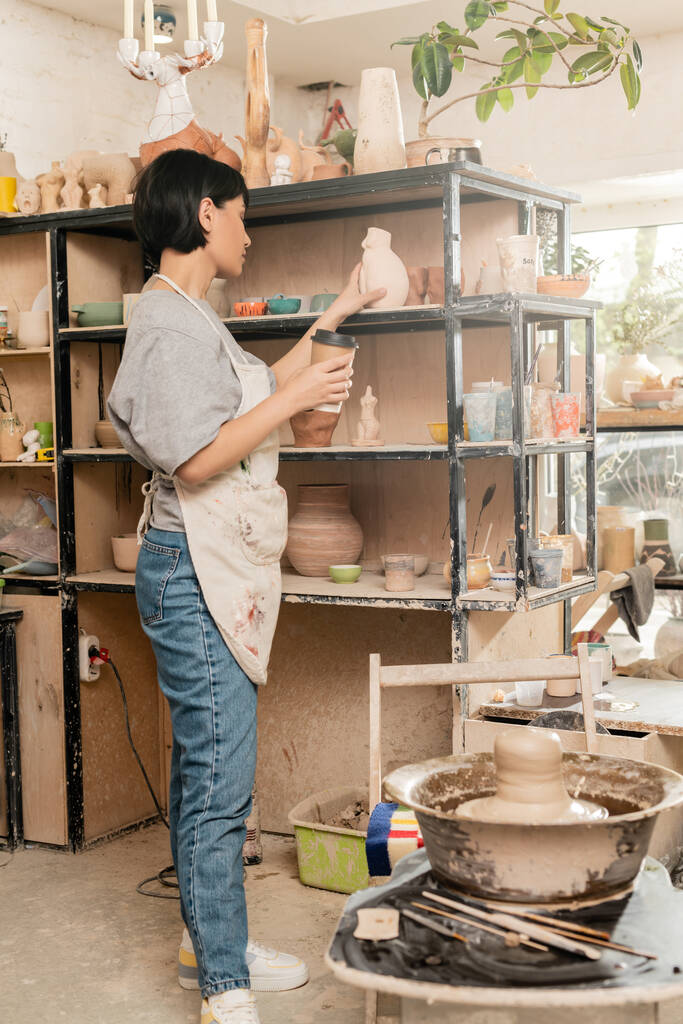 Молода азіатка-художниця в фартусі тримає каву і скульптуру біля глини на гончарному колесі і стійку в керамічній майстерні на фоні, майстер створює унікальні керамічні деталі - Фото, зображення