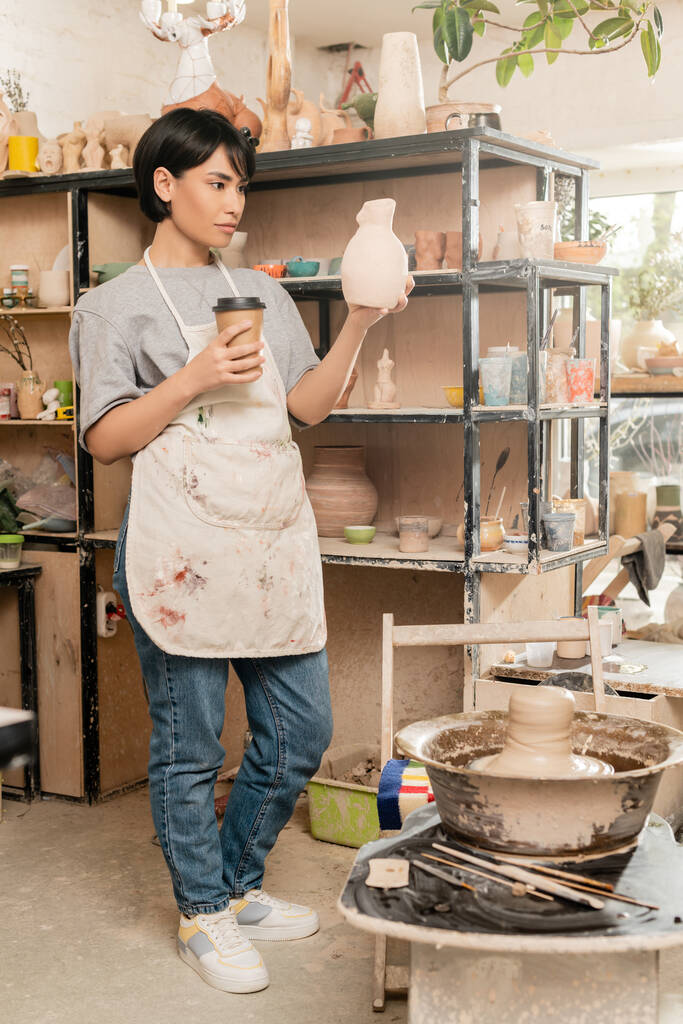 Young asian female artist in apron holding sculpture and coffee to go near clay on pottery wheel, tools and rack in ceramic workshop, artisan creating unique pottery pieces - Photo, Image