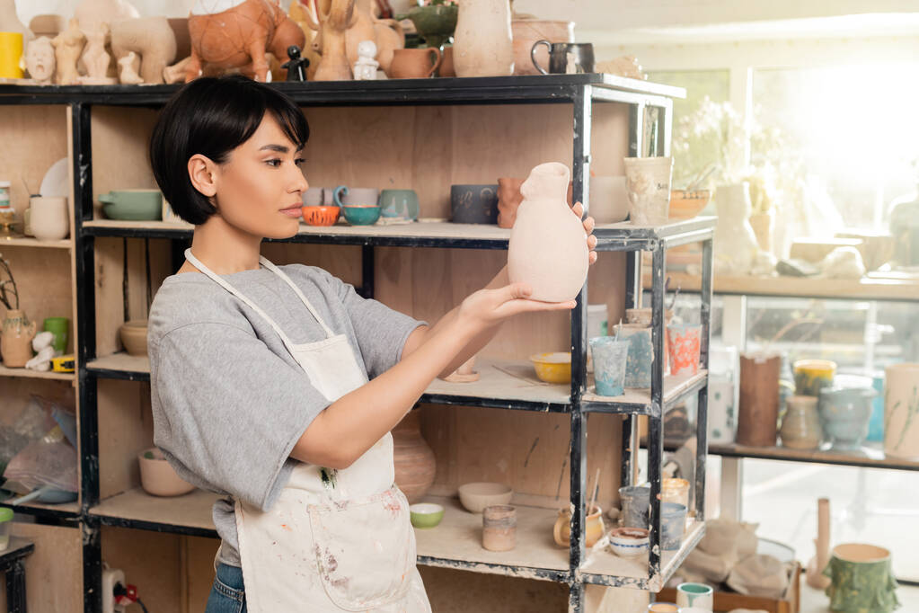 若いブルネットのアジアの女性の職人は日没時に背景にぼやけたセラミックワークショップで棚とラックの近くに粘土彫刻を保持エプロン、職人はユニークな陶器作品を作成します - 写真・画像