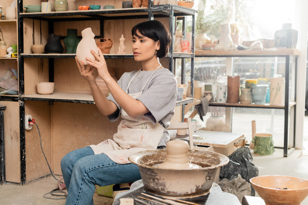 Молодая азиатская художница в фартуке, держащая керамическую скульптуру, сидя рядом с глиной на гончарном колесе и инструментах в размытой студии на закате, концепция процесса скульптуры из глины - Фото, изображение