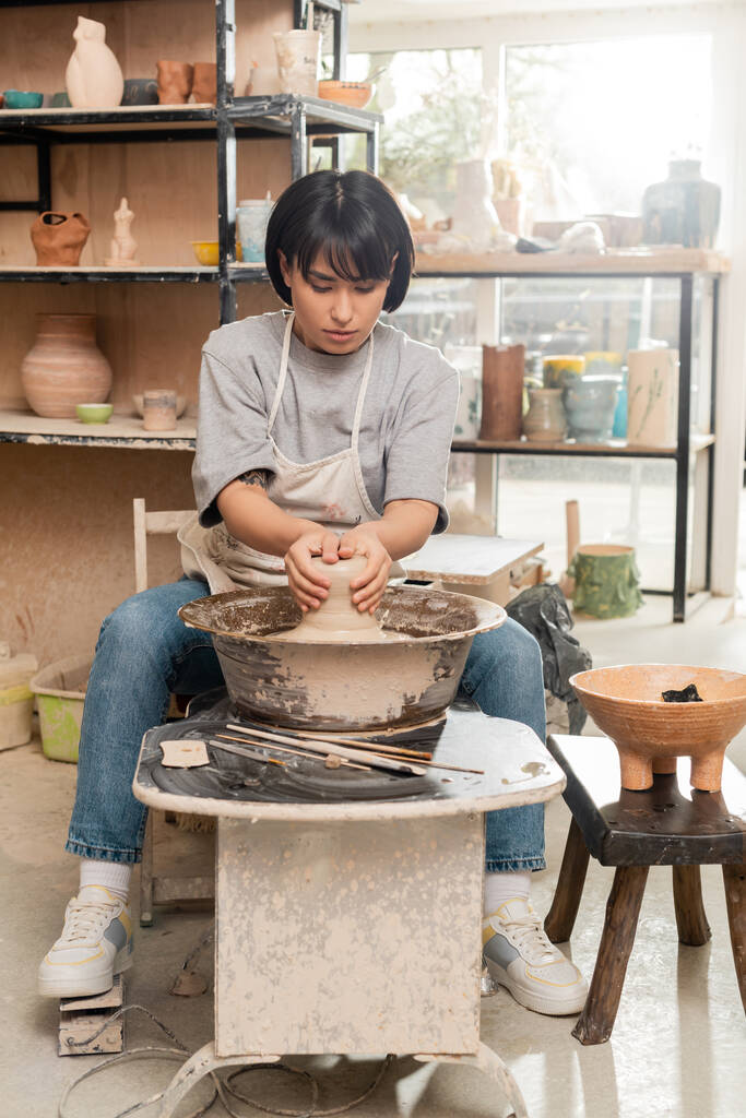 Mladá asijská žena hrnčíř v zástěře lití mokrý jíl na keramice kolo v blízkosti nástroje a miska s houbou při práci v keramické studio na pozadí, hliněné sochařství proces koncepce - Fotografie, Obrázek
