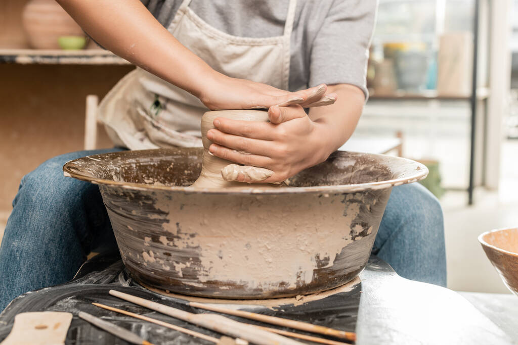 Cropped view of blurred female artist in apron shaping wet clay and working on pottery wheel near wooden tools on table in ceramic workshop, clay sculpting process concept - Photo, Image