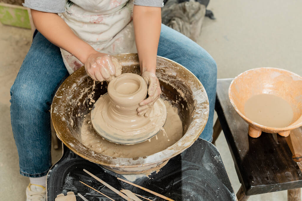 Vista de alto ângulo do oleiro fêmea jovem no avental derramando água na argila e roda de cerâmica girando perto tigela e ferramentas na oficina de arte, cerâmica qualificada que faz o conceito - Foto, Imagem