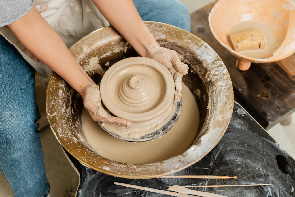 Vue du dessus de jeune artisan en tablier faisant forme d'argile humide sur la roue de poterie près du bol avec éponge et outils sur la table en atelier d'art, concept de fabrication de poterie habile - Photo, image