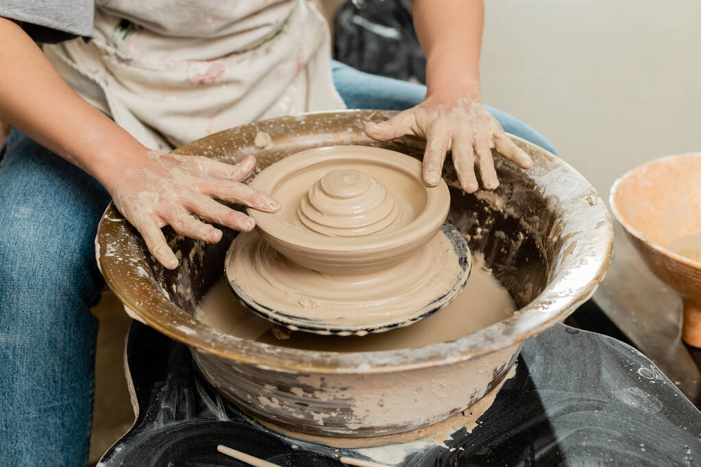 Vista ritagliata di giovane artigiana in grembiule che fa forma di argilla sulla ruota della ceramica di filatura in laboratorio di ceramica sfocata sullo sfondo, concetto di ceramica qualificato - Foto, immagini