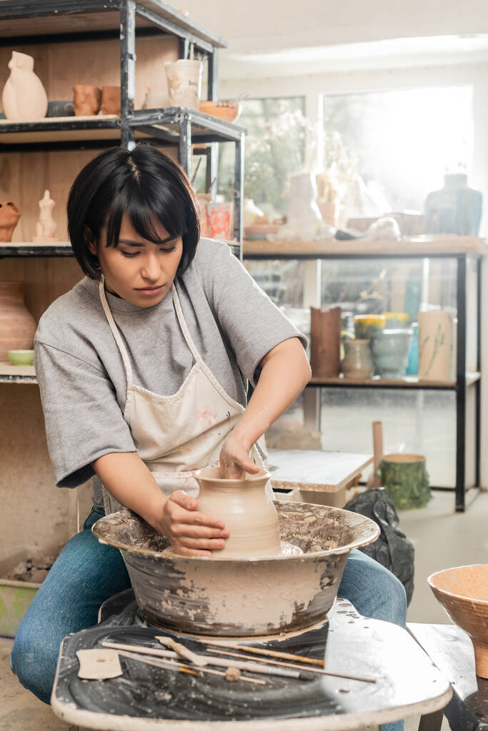 Young brunette asian female artisan in apron shaping clay vase while working on pottery wheel near wooden tools and bowl in blurred ceramic workshop, pottery creation process - Photo, Image