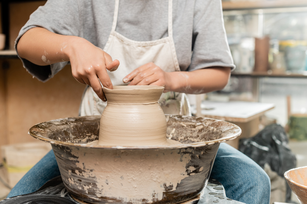 Vista recortada de la joven ceramista en delantal y ropa de trabajo haciendo forma de jarrón de arcilla y trabajando con rueda de cerámica en taller de cerámica borrosa, proceso de creación de cerámica - Foto, Imagen