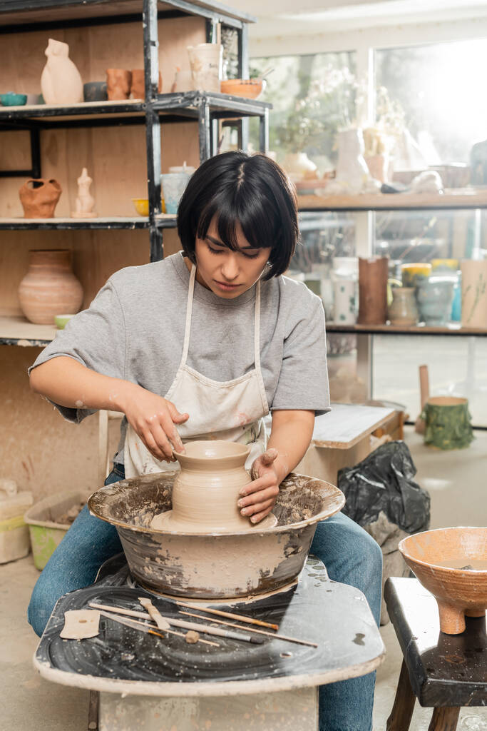 Jeune artisan asiatique brune en tablier et vêtements de travail faisant vase d'argile et travaillant sur la rotation de la roue de poterie dans un atelier de céramique floue à l'arrière-plan, processus de création de poterie - Photo, image