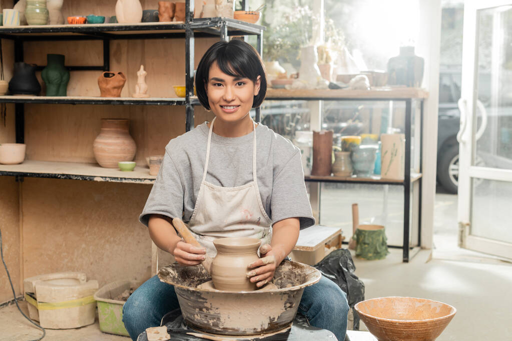 Usmívající se mladá brunetka asijské žena řemeslník v zástěře při pohledu na kameru, zatímco drží dřevěný nástroj v blízkosti jílu na keramické kolo v keramické studio na pozadí, jílu tvarování a tváření procesu - Fotografie, Obrázek