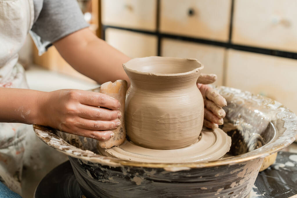 Vista ritagliata di giovane donna artigiana in grembiule rendendo vaso di argilla con raschietto di legno su ruota ceramica in laboratorio di ceramica sfocata, modellatura argilla e processo di formazione - Foto, immagini