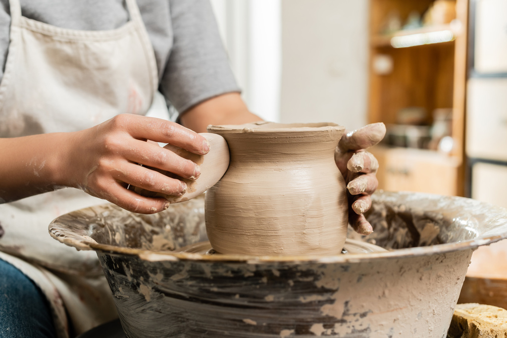 Обрізаний вид розмитого молодого майстра в фартусі, який тримає дерев'яний скребок, роблячи глиняну вазу на керамічному колесі в художній студії на фоні, формування глини і процес формування - Фото, зображення
