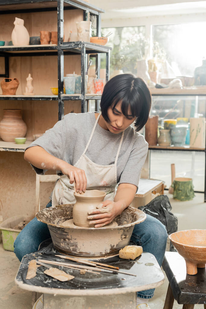 Young brunette asian craftswoman in apron making shape of clay vase while working with pottery wheel near sponge and wooden tools in ceramic workshop, clay shaping and forming process - Photo, Image
