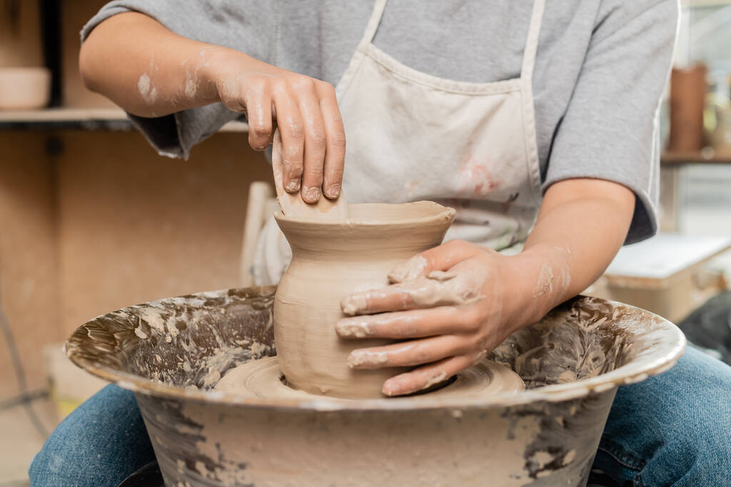 Oříznutý pohled na mladé řemeslnice v zástěře při výrobě holení hliněné vázy s dřevěným nářadím při práci s točícím se keramickým kolem v keramické dílně, tvarování a tváření hlíny - Fotografie, Obrázek