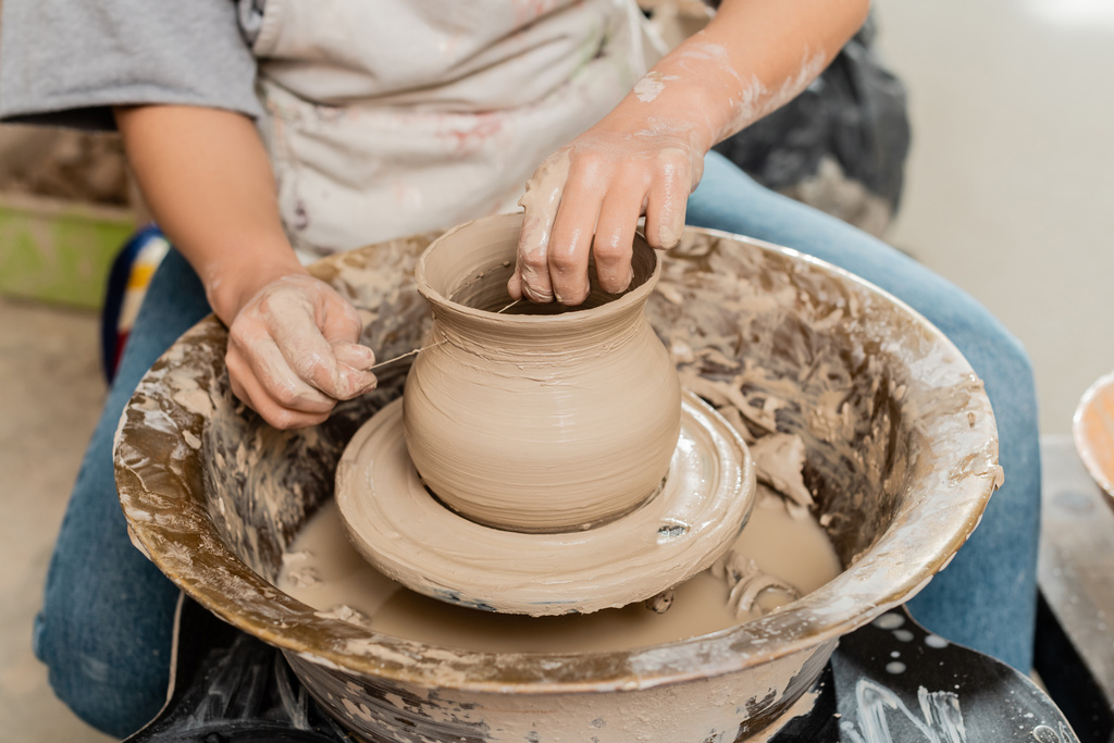 Vista cortada do artesão feminino morena em vaso de argila de corte de avental na roda de cerâmica enquanto trabalhava em oficina de cerâmica no fundo, modelagem e processo de formação de argila - Foto, Imagem