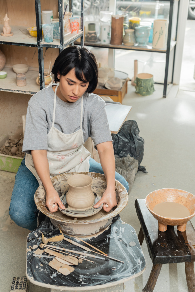 Joven ceramista asiática en delantal moldeando jarrón de arcilla con herramientas en la rueda de cerámica giratoria cerca de esponja y tazón con agua en estudio de cerámica, producción y proceso de cerámica artesanal - Foto, imagen
