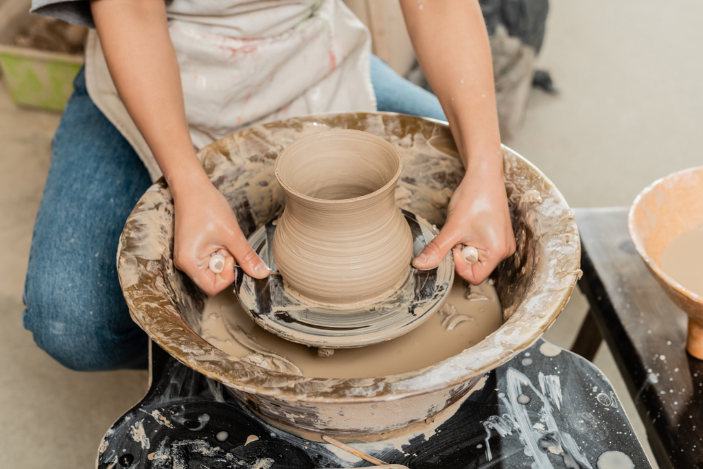陶磁器工房の背景に水を入れたボールの近くに回転する陶器製の車輪の上のエプロン切断粘土花瓶の若い女性職人の作物ビュー、職人の陶器の生産とプロセス - 写真・画像