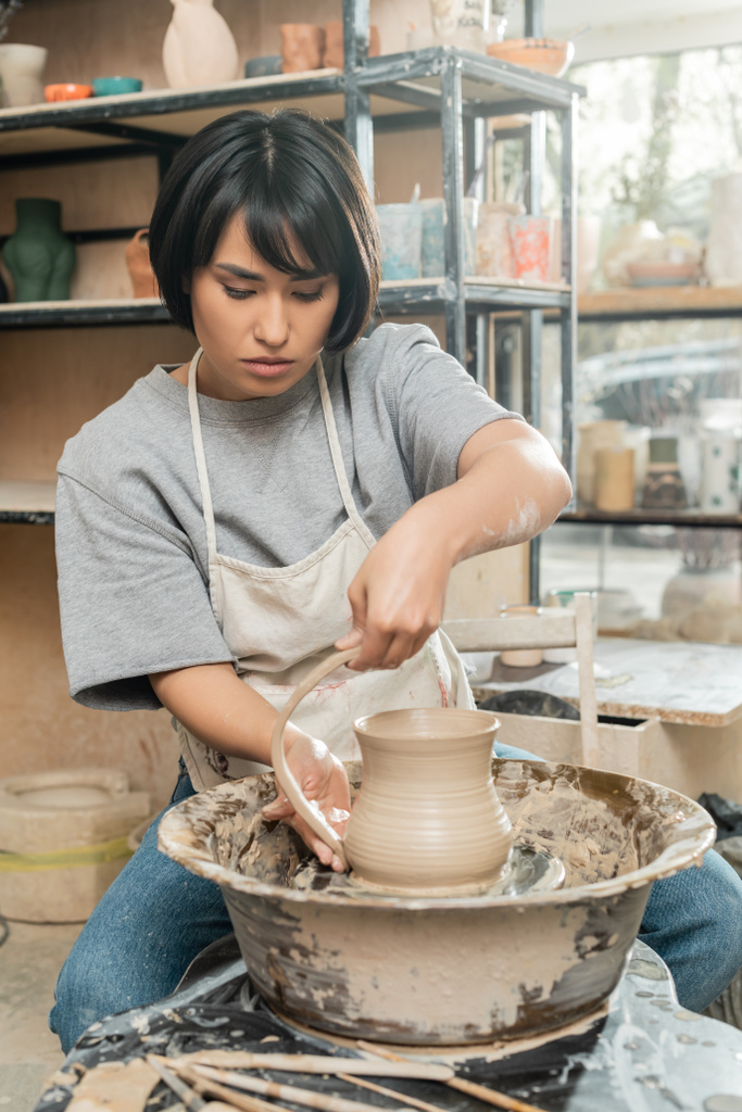 Joven morena asiática artesana en taller haciendo jarra de arcilla mientras trabaja en hilar rueda de cerámica cerca de herramientas de madera borrosas en taller de cerámica, producción y proceso de cerámica artesanal - Foto, Imagen