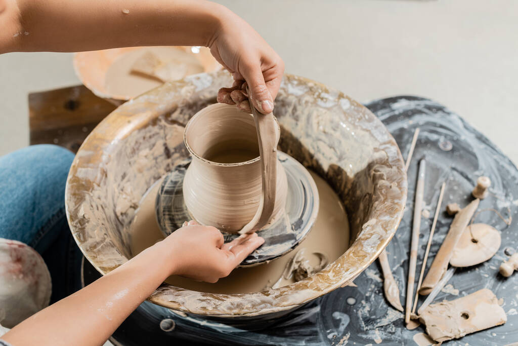 Vista de alto ângulo de oleiro feminino jovem em roupas de trabalho criando jarro de barro e trabalhando com roda de cerâmica perto de ferramentas de madeira na mesa no estúdio de arte, produção e processo de cerâmica artesanal - Foto, Imagem