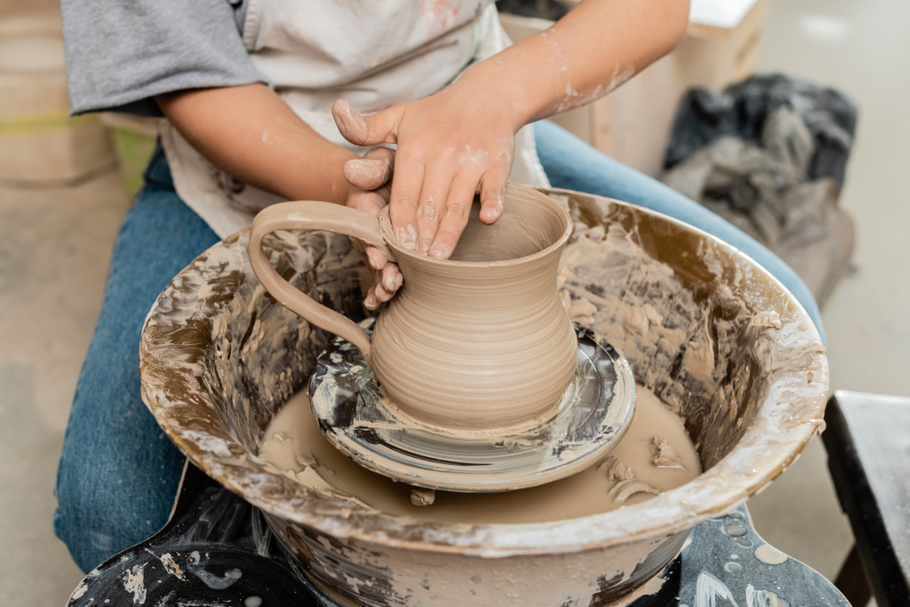 陶芸ワークショップ、職人技の陶器の生産とプロセスで作業しながら、テーブルの上に陶器の車輪の上に粘土のジャグを作成エプロンで若い女性アーティストの作物のビュー - 写真・画像