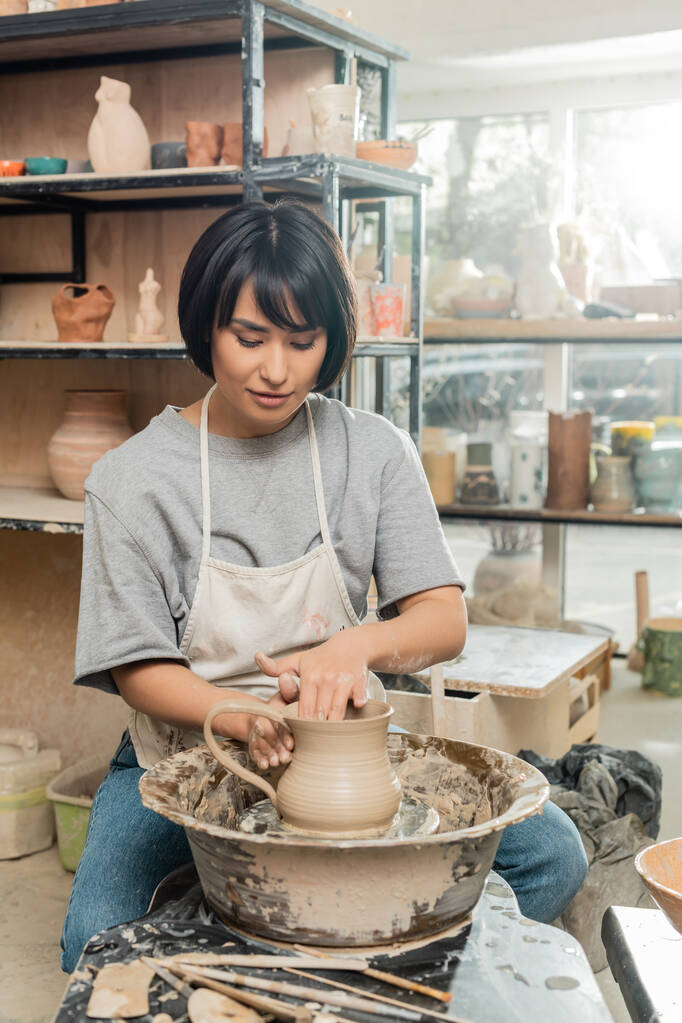 Jovem asiática artista feminina em avental e workwear criando jarro de barro na roda de cerâmica perto de ferramentas de madeira na mesa em oficina de cerâmica borrada no fundo, técnica de modelagem de barro e processo - Foto, Imagem