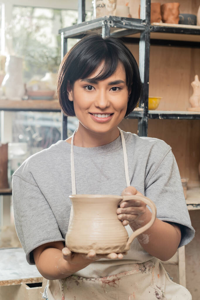 Önlüklü, iş kıyafetli, killi sürahi tutan ve arka plandaki bulanık seramik atölyesinde kameraya bakan Asyalı genç esmer kadın zanaatkarın portresi, kil şekillendirme tekniği ve işleme süreci - Fotoğraf, Görsel