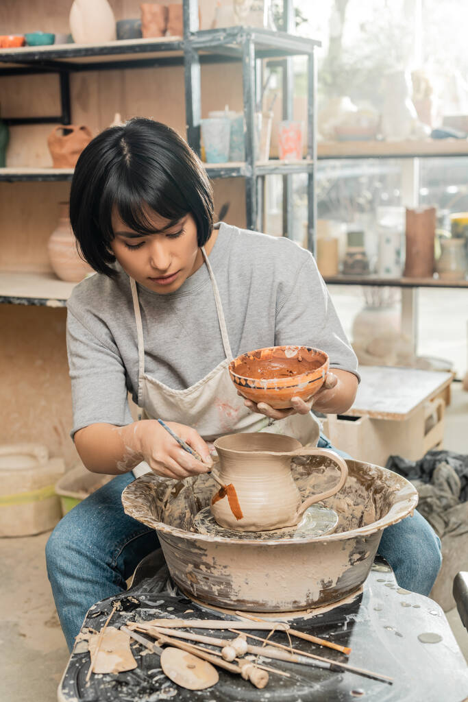 Giovane bruna asiatica artigiana in grembiule pittura su brocca di argilla sulla ruota della ceramica mentre si lavora vicino a strumenti di legno sul tavolo in laboratorio di ceramica sullo sfondo, tecnica di modellatura dell'argilla e processo - Foto, immagini