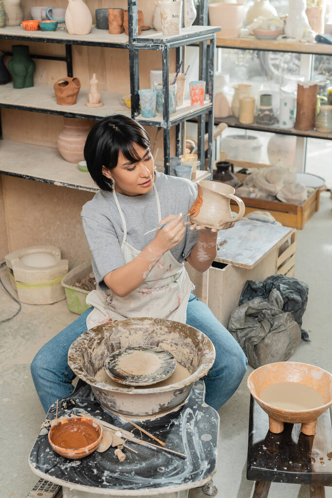 Високий кут зору молодої азіатки-художниці в фартуховому живописі на глиняному глечику біля гончарного колеса, дерев'яних інструментів і чаші з водою в керамічній майстерні, техніка формування глини і процес - Фото, зображення