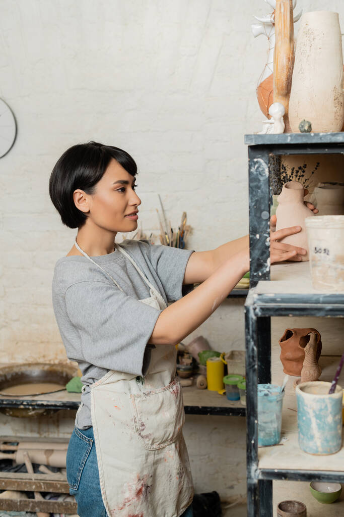Smiling young asian craftswoman in apron putting ceramic sculpture on shelf on rack while standing in blurred art workshop at background, clay shaping technique and process - Photo, Image