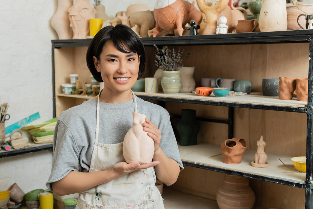 Porträt eines fröhlichen jungen asiatischen Kunsthandwerkers in Schürze mit Keramikskulptur und Blick auf die Kamera in der Nähe von Gestell in verschwommener Kunstwerkstatt im Hintergrund, Töpferatelier-Szene - Foto, Bild